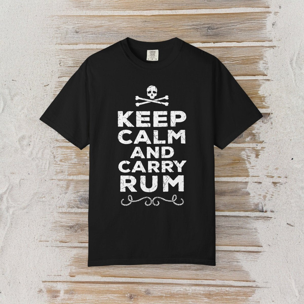 A black short-sleeve crewneck t-shirt with a white graphic print reading 'KEEP CALM AND CARRY RUM' below a skull and crossbones, displayed flat against a rustic light wood plank background.