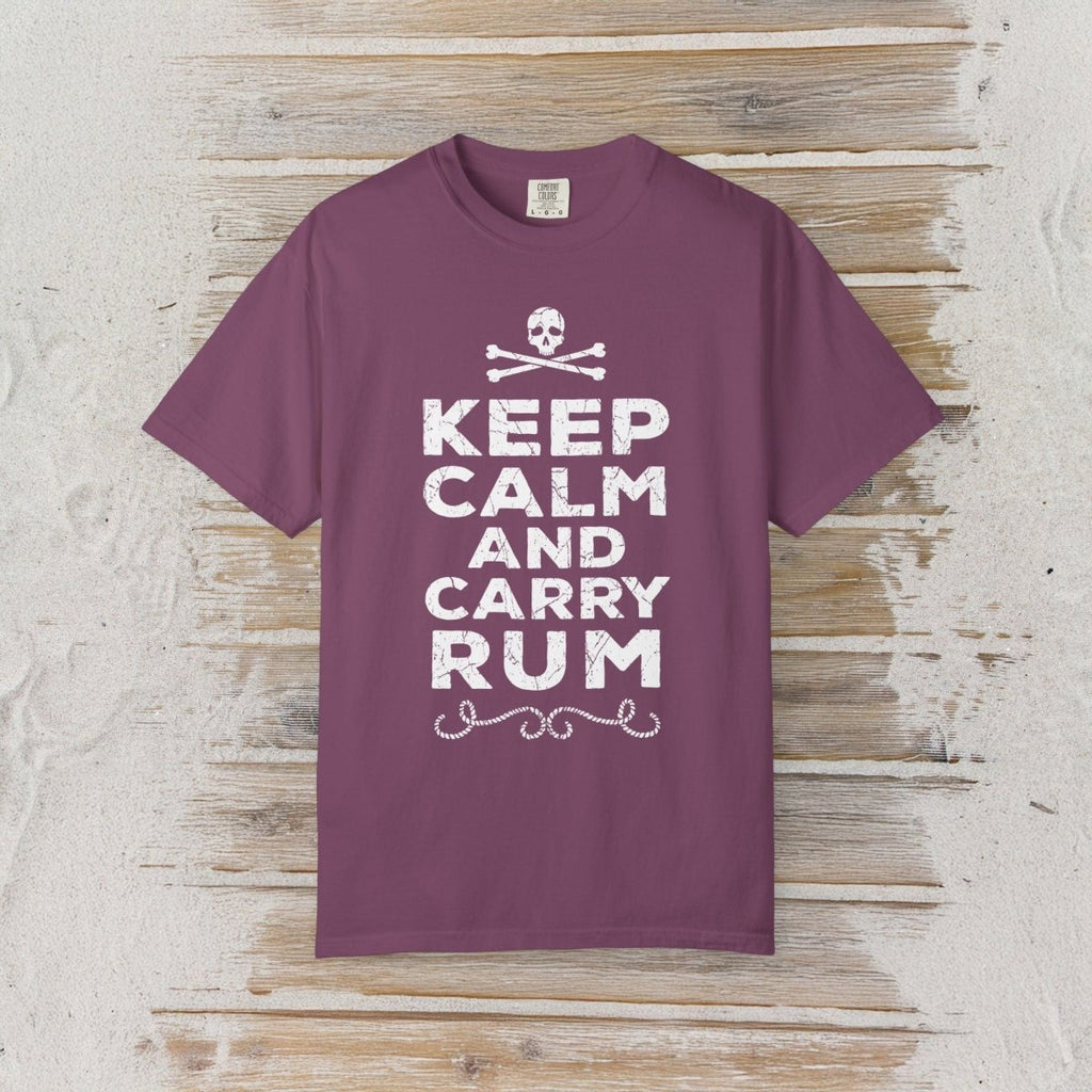 A purple short-sleeve t-shirt with a white graphic print reading 'KEEP CALM AND CARRY RUM' with a skull and crossbones above, displayed flat on a rustic wooden plank background.