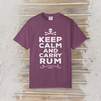 A purple short-sleeve t-shirt with a white graphic print reading 'KEEP CALM AND CARRY RUM' with a skull and crossbones above, displayed flat on a rustic wooden plank background.