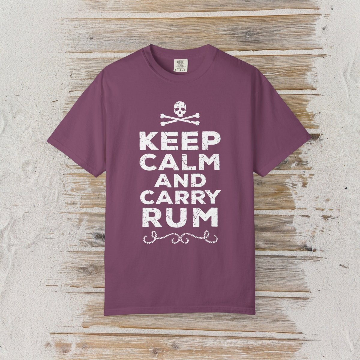 A purple short-sleeve t-shirt with a white graphic print reading 'KEEP CALM AND CARRY RUM' with a skull and crossbones above, displayed flat on a rustic wooden plank background.
