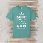 A teal short-sleeve t-shirt with a white graphic print reading 'KEEP CALM AND CARRY RUM' below a skull and crossbones, displayed flat on a rustic light wood background.