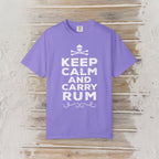 A purple short-sleeve crewneck t-shirt with a white graphic print reading 'KEEP CALM AND CARRY RUM' below a skull and crossbones, displayed flat on a rustic light wood plank background.