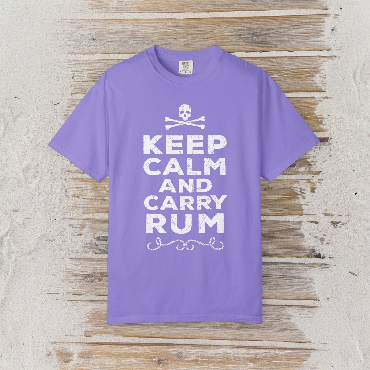 A purple short-sleeve crewneck t-shirt with a white graphic print reading 'KEEP CALM AND CARRY RUM' below a skull and crossbones, displayed flat on a rustic light wood plank background.