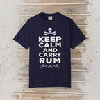 A navy blue short-sleeve t-shirt with a white graphic print reading 'KEEP CALM AND CARRY RUM' below a skull and crossbones, displayed flat against a rustic light wood plank background.