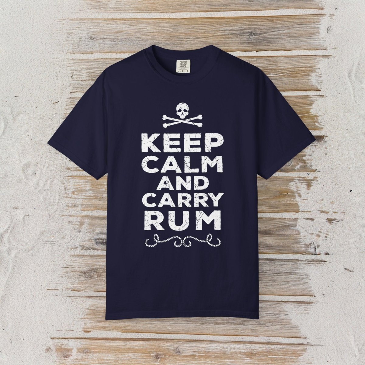 A navy blue short-sleeve t-shirt with a white graphic print reading 'KEEP CALM AND CARRY RUM' below a skull and crossbones, displayed flat against a rustic light wood plank background.
