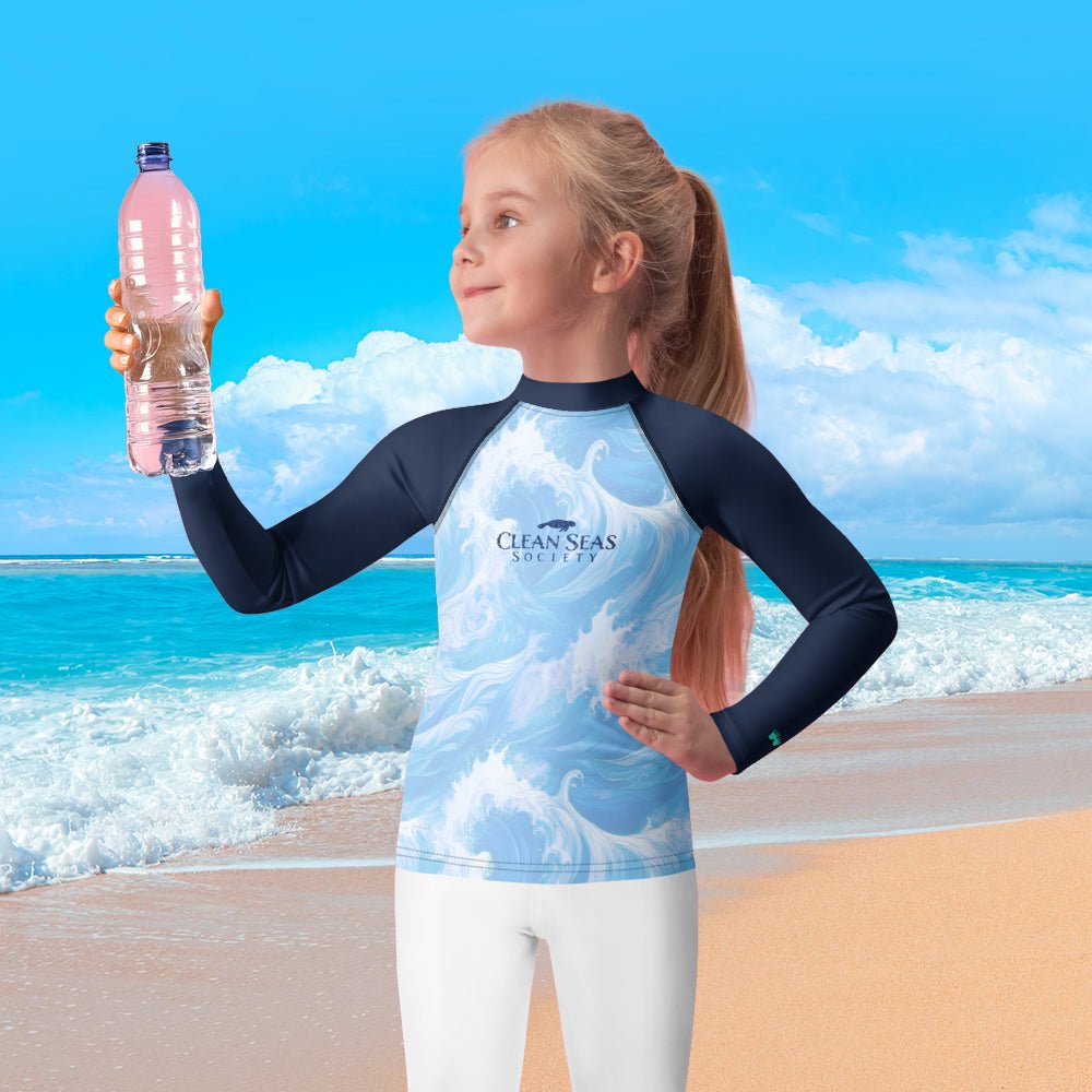 Child wearing a 'Clean Seas Society' rash guard shirt on a beach holding a water bottle.