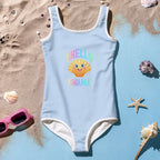 Girl's One Piece Sky Blue Swimsuit with cute cartoon shell saying "SHello Gwama' " lying in beach sand next to seashells and sunglasses