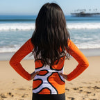 A tween girl with long dark hair stands on a sandy beach facing the ocean, wearing a long-sleeve athletic top with an orange, white, and black abstract pattern.