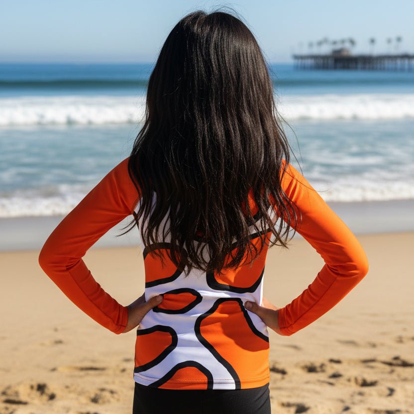 A tween girl with long dark hair stands on a sandy beach facing the ocean, wearing a long-sleeve athletic top with an orange, white, and black abstract pattern.