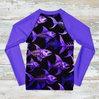 A long-sleeve raglan rash guard shirt featuring a black front panel with a repeating purple and blue fish skeleton pattern, paired with solid purple sleeves and collar, laid flat on a distressed white wood background.