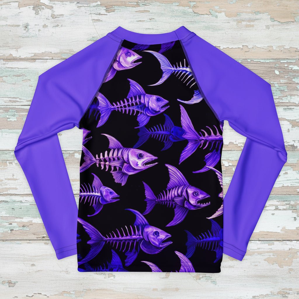 A long-sleeve raglan rash guard shirt featuring a black front panel with a repeating purple and blue fish skeleton pattern, paired with solid purple sleeves and collar, laid flat on a distressed white wood background.