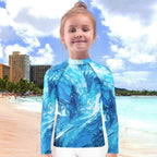 Child wearing a blue wave patterned shirt on a beach with buildings and ocean in the background