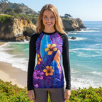 Woman wearing a colorful beach rashie with a surfboard design standing in front of green foliage and La Jolla Cove in the background