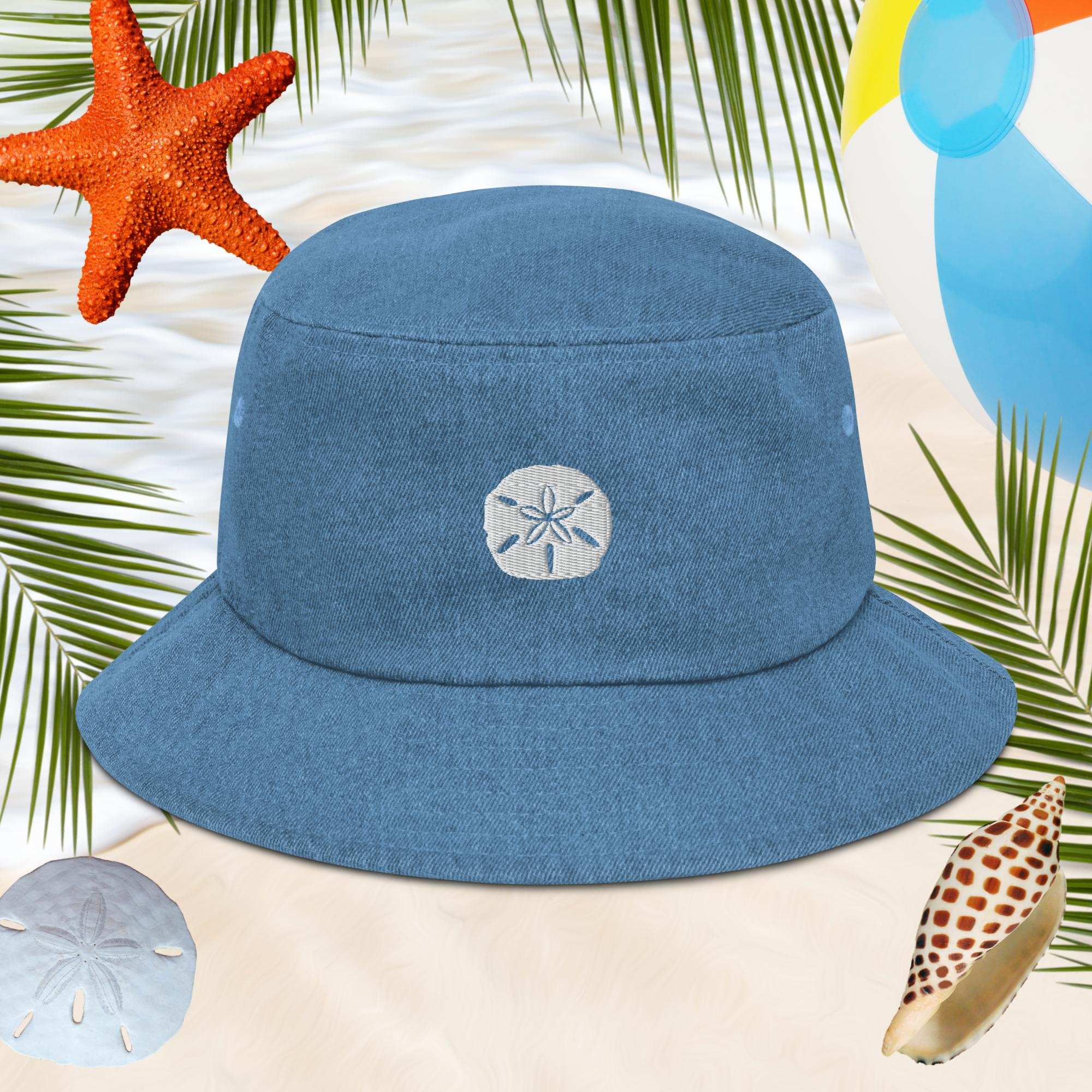 Blue bucket hat with an embroidered sand dollar design on a beach-themed background with starfish, shells, and palm leaves.