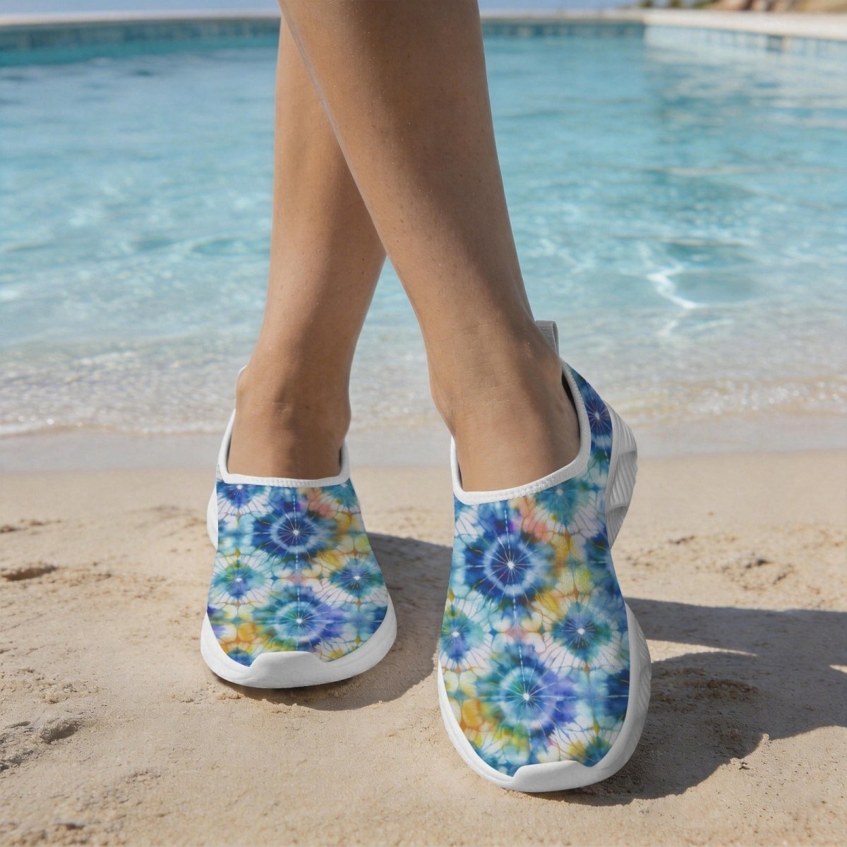 A person standing on a sandy beach by a swimming pool wearing Women's Cute Water Shoes with a blue, yellow, and white shibori-tie-dye pattern.