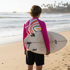 A teen dude seen from behind on Laguna Beach, wearing a bright pink long-sleeve rash guard with a colorful abstract wave pattern on the back and holding a white surfboard.