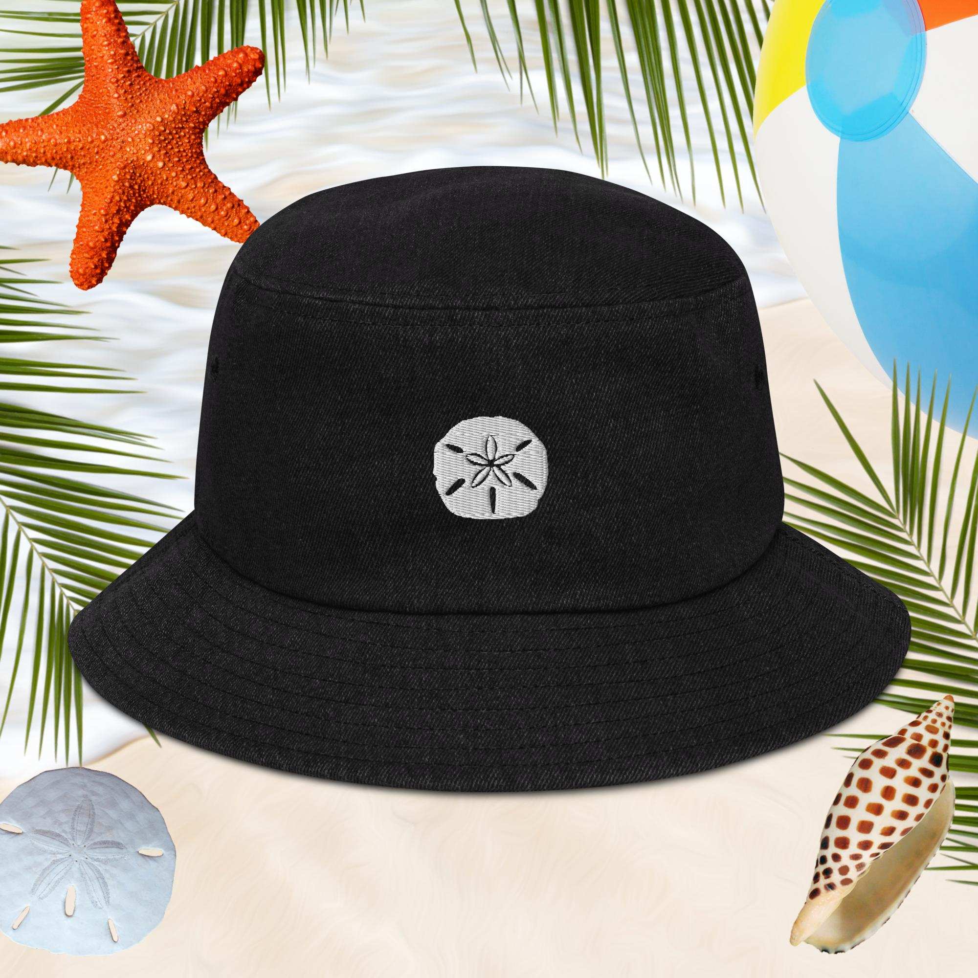 Black bucket hat with embroidered sand dollar design on a beach-themed background with starfish, shells, and balloons.