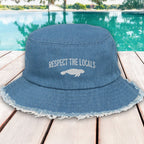 Blue bucket hat with 'Respect the Locals' text and manatee graphic on a wooden deck by a pool.