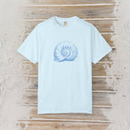 A light blue short-sleeve crewneck t-shirt featuring a blue nautilus shell graphic on the chest, laid flat on a rustic wooden plank background.