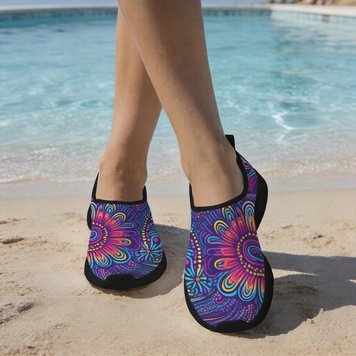 A person standing on a sandy beach by a swimming pool wearing slip-on water shoes with a vibrant purple, pink, and blue island tribal pattern and black elastic trim.