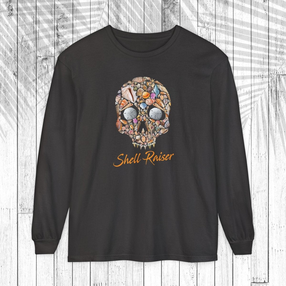 A black long-sleeve t-shirt with a colorful skull graphic made of various small objects and the text 'Shell-Raiser' in orange cursive, displayed on a white wood plank background.