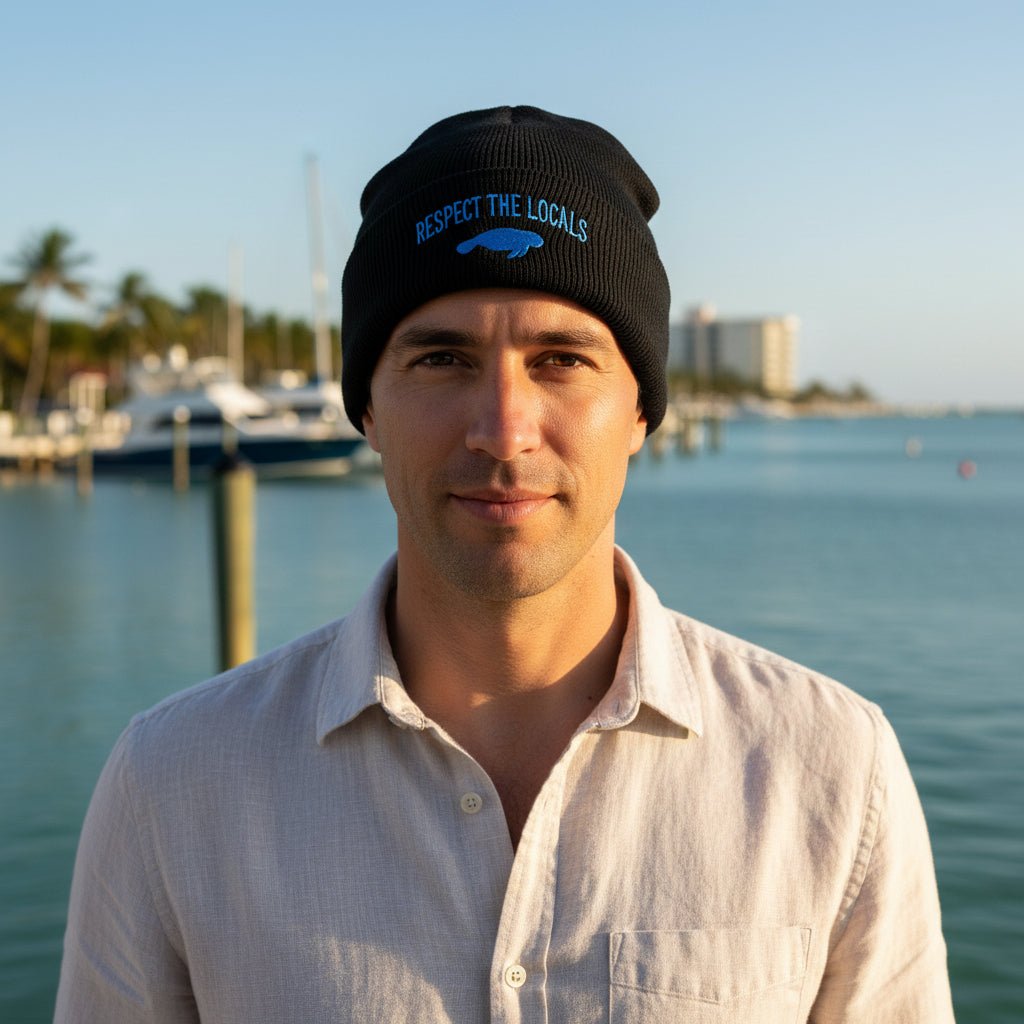 A black knit beanie with a folded cuff featuring the text 'RESPECT THE LOCALS' in blue and a blue manatee silhouette below it, worn by a pontoon boat captain at manatee cove