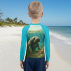 A child standing on a beach, viewed from behind, wearing a long-sleeve swim shirt with bright turquoise sleeves and a large printed underwater scene on the back featuring a manatee.