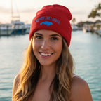 Woman wearing a red knit Save The Manatees beanie hat with a folded cuff featuring the text 'RESPECT THE LOCALS' in blue and a blue manatee graphic below it, standing on a boat on Manatee River