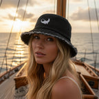 Woman wearing a dark navy denim bucket hat with a frayed, distressed edge and a white embroidered stylized manta ray on the front. 