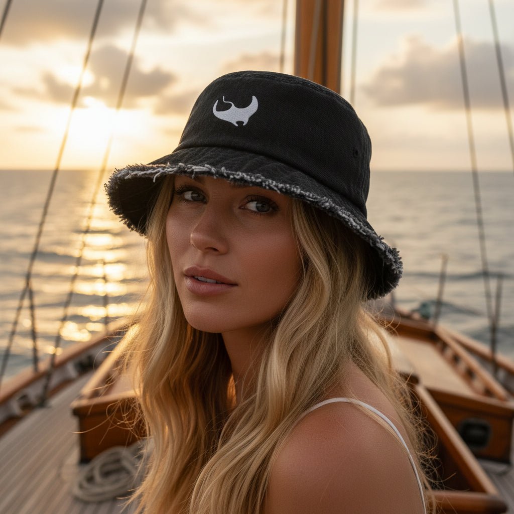 Woman wearing a dark navy denim bucket hat with a frayed, distressed edge and a white embroidered stylized manta ray on the front. 