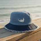A blue denim bucket hat with a dark navy blue frayed brim and a white embroidered Manta Ray logo on the front, sitting on a beachside boardwalk