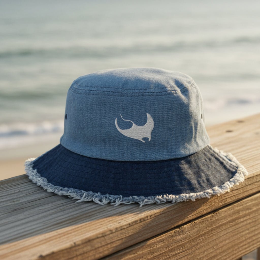 A blue denim bucket hat with a dark navy blue frayed brim and a white embroidered Manta Ray logo on the front, sitting on a beachside boardwalk