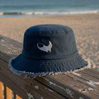 Navy blue bucket hat with a white manta ray embroidery on a beachside boardwalk