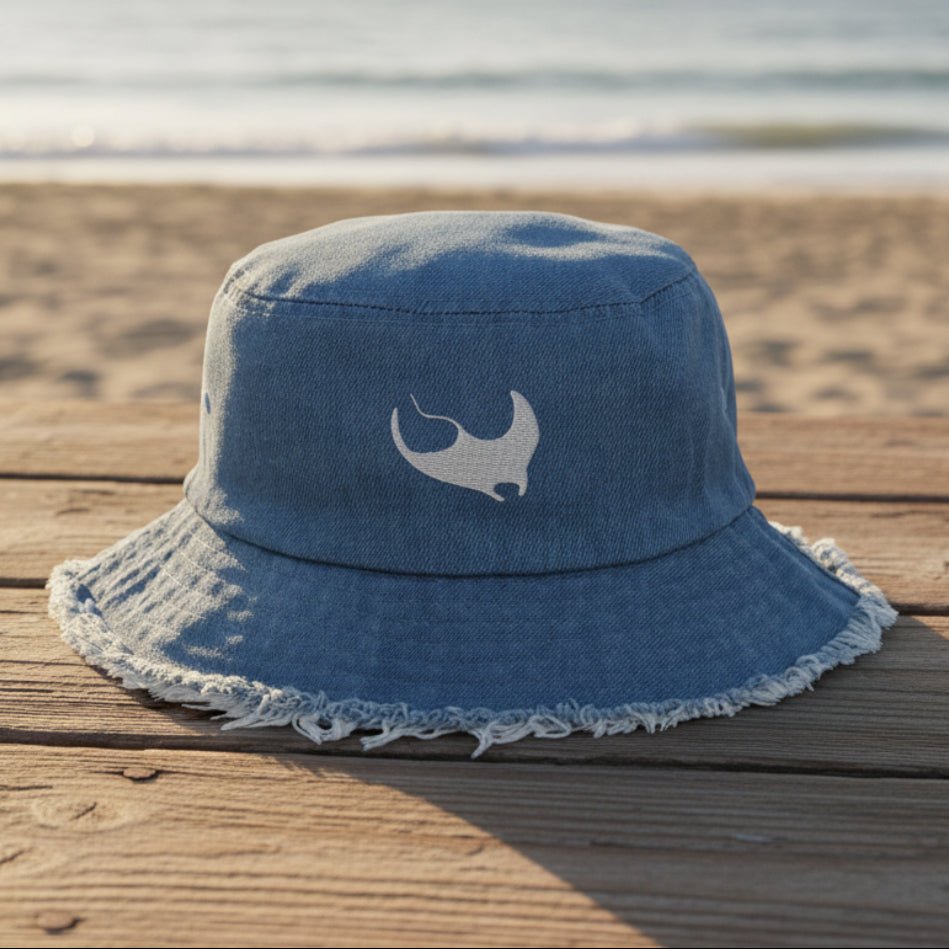 A light blue denim bucket hat with a frayed edge and a white embroidered stylized Manta Ray embroidery on the front, shown on a beachside boardwalk