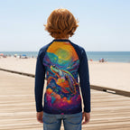 Kid in a long-sleeve raglan rash guard with a dark navy blue base, featuring a large, vibrant, multicolor sea turtle print, standing on the Ocean City Boardwalk 