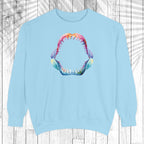 A light blue crewneck sweatshirt featuring a colorful graphic of an open shark mouth with sharp teeth on the chest, displayed against a white wood plank background with a leaf shadow overlay.