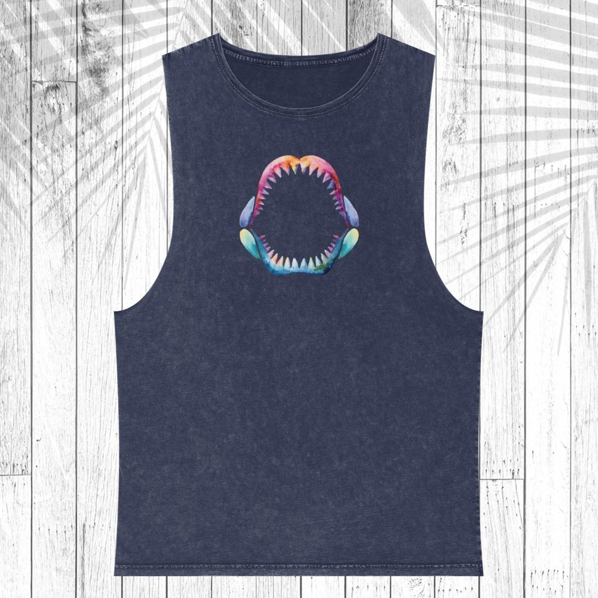 A sleeveless dark navy tank top with a colorful shark mouth graphic on the chest, displayed flat against a white wood plank background with a palm leaf overlay.
