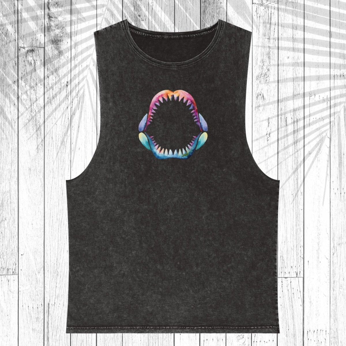 A black washed sleeveless tank top featuring a colorful graphic of an open shark mouth on the chest, displayed flat against a white wood plank background with a palm leaf overlay.