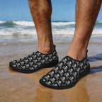 Black slip-on water shoes with a grey skull and crossbones pattern, worn on a person's feet at the beach with ocean waves in the background.