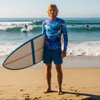 Dude wearing colorful men's Swim Shirt holding a surfboard on a beach with ocean waves in the background