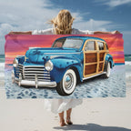 Woman on Moonlight Beach holding up a rectangular beach towel featuring a vintage blue wood-paneled station wagon on a sandy beach under a colorful sunset sky.