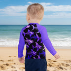 A toddler viewed from the back wearing a long-sleeve rash guard with purple sleeves and a black back featuring a purple fish skeleton pattern, set against a myrtlebeach background.