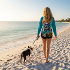 A woman on Naples Beach walks barefoot with a Boston Terrier on a leash, wearing a teal long-sleeve swim top with a large French bulldog graphic on the front and black swim shorts.