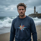 A dark heather gray crewneck sweatshirt featuring a colorful compass rose graphic on the chest, worn by a guy walking in a northern shore beach near a lighthouse 