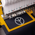 Close-up of an embroidery machine stitching a white rope-style peace sign design onto dark navy fabric inside a yellow hoop.