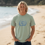 A smiling blonde man standing on a sandy beach wearing a light sage green t-shirt with a blue nautilus shell graphic on the chest, paired with dark blue jeans.