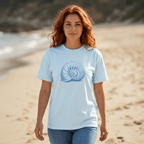 A woman with red hair stands on a sandy beach wearing a light blue crewneck t-shirt with a blue nautilus seashell graphic on the front, paired with blue jeans.