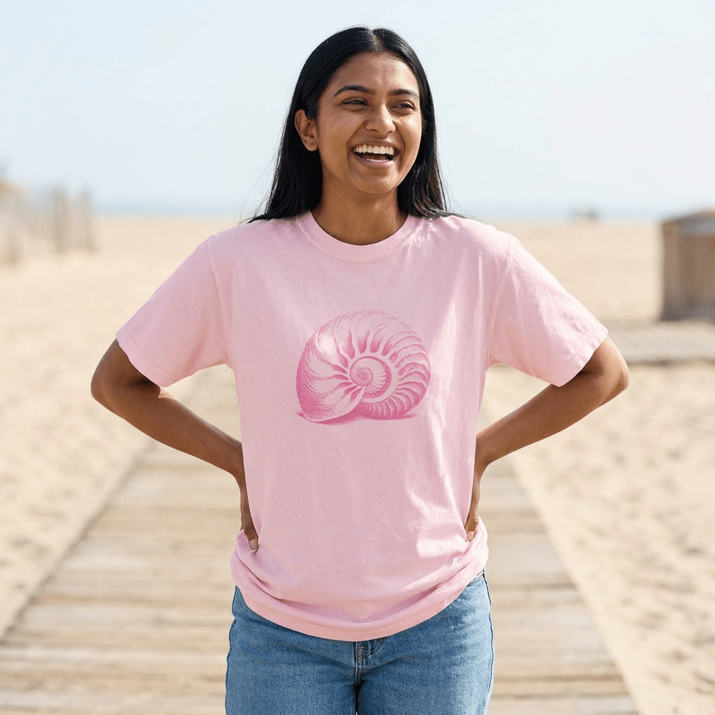 A woman smiling on a wooden boardwalk at the beach, wearing a light pink crewneck t-shirt with a pink nautilus shell graphic on the front, paired with blue jeans.