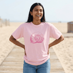 A woman smiling on a wooden boardwalk at the beach, wearing a light pink crewneck t-shirt with a pink nautilus shell graphic on the front, paired with blue jeans.
