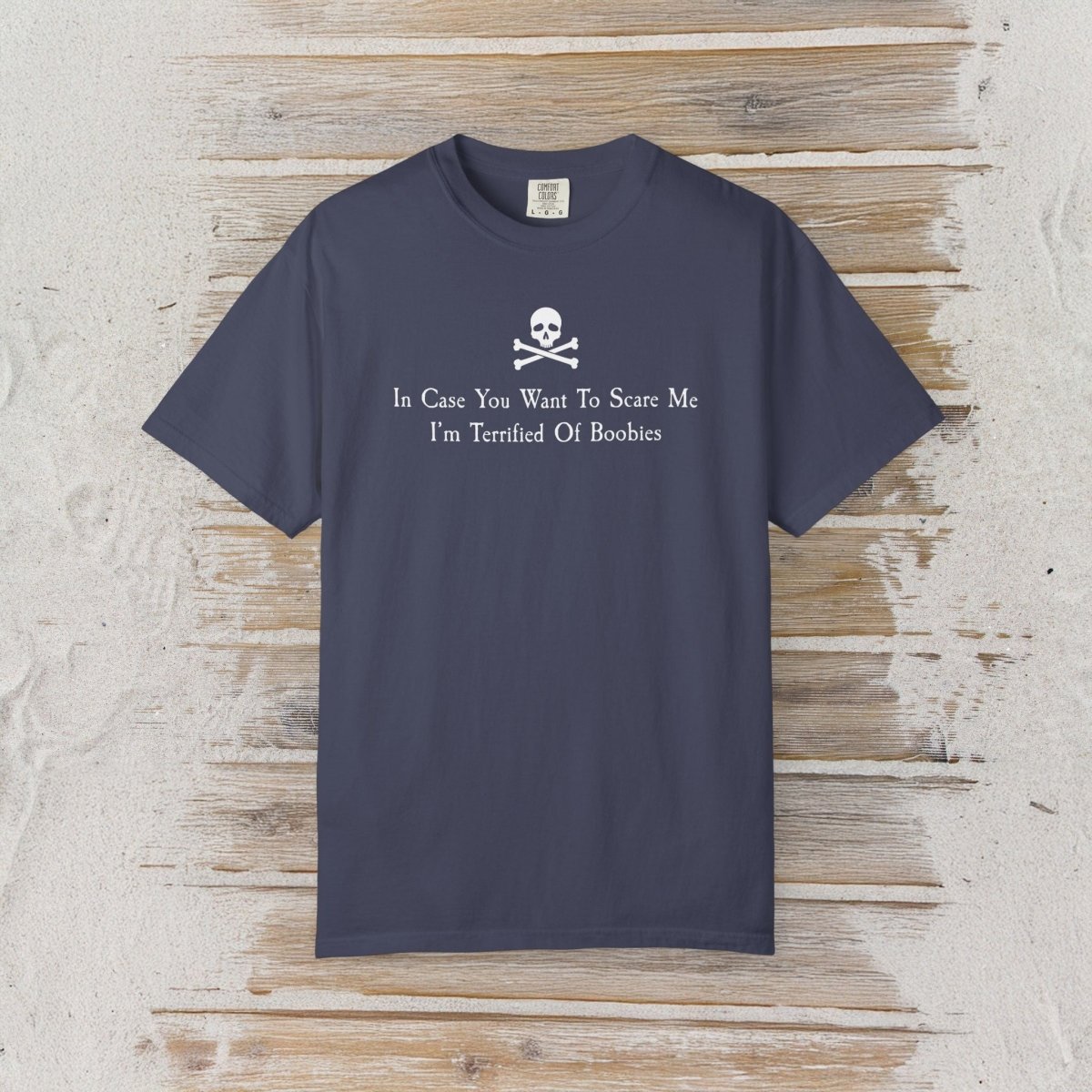 A navy blue short-sleeve t-shirt with a white skull and crossbones graphic and the text 'In Case You Want To Scare Me I'm Terrified Of Boobies' printed on the chest, displayed flat on a rustic light wood background.